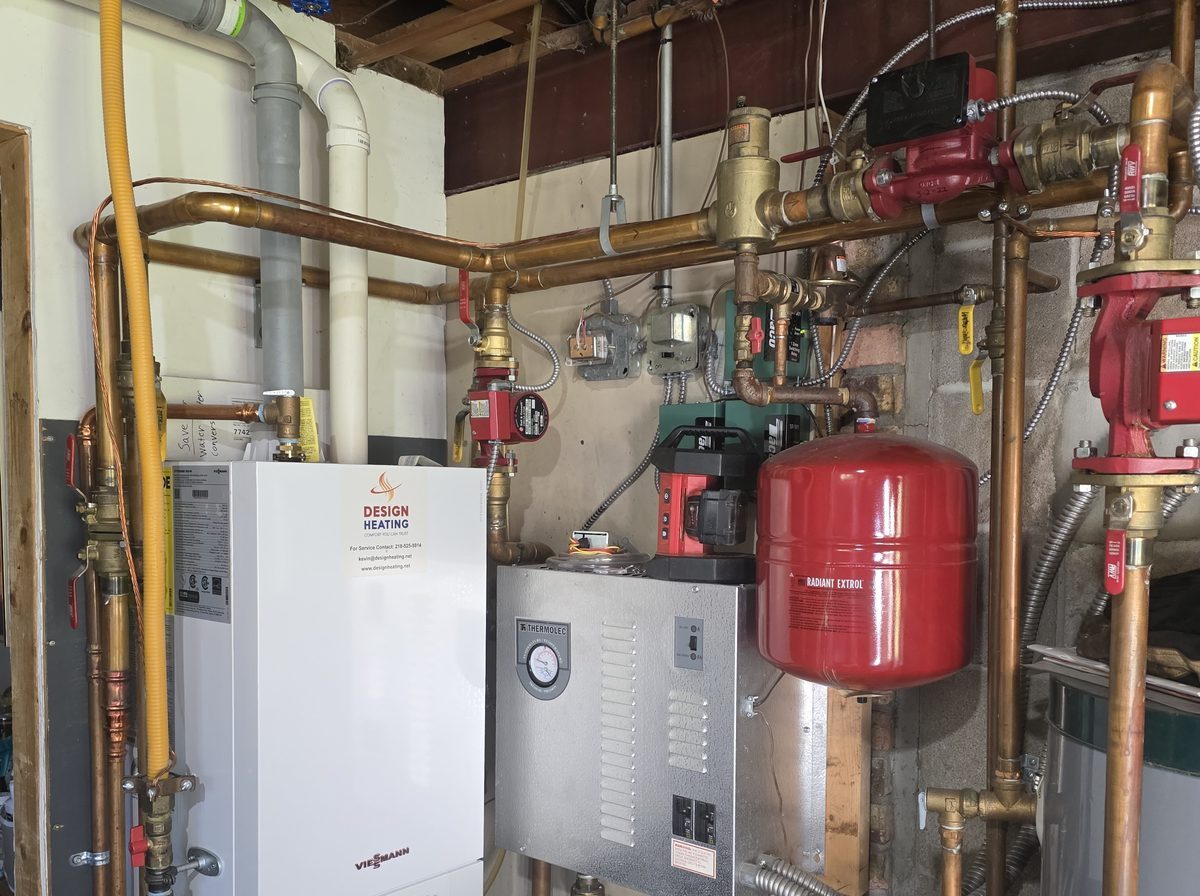 Wall-hung Viessmann gas boiler with a Design Heating service sticker, next to an electric boiler, red expansion tank and copper piping in a basement mechanical room.